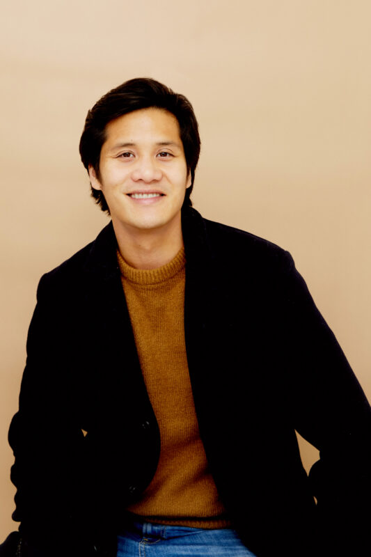 A headshot of Tam Lan Truong. He is wearing a brown shirt, a black blazer and is smiling.